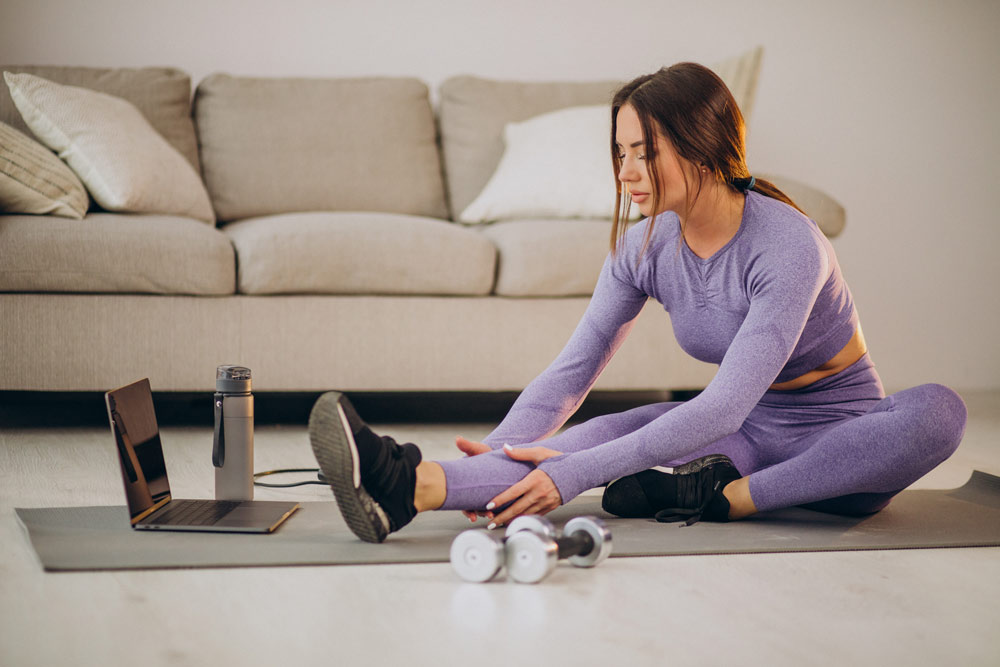 A fit woman on an exercise floormat stretching and looking at a laptop with dumbbells next to her.