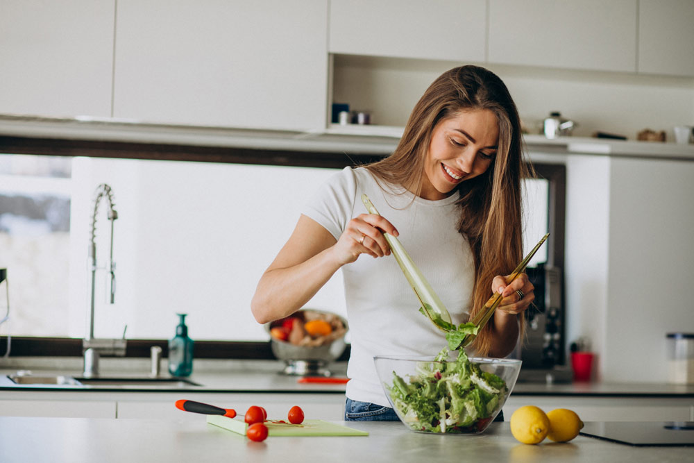 A smiling woman in a kitchen making a salad.